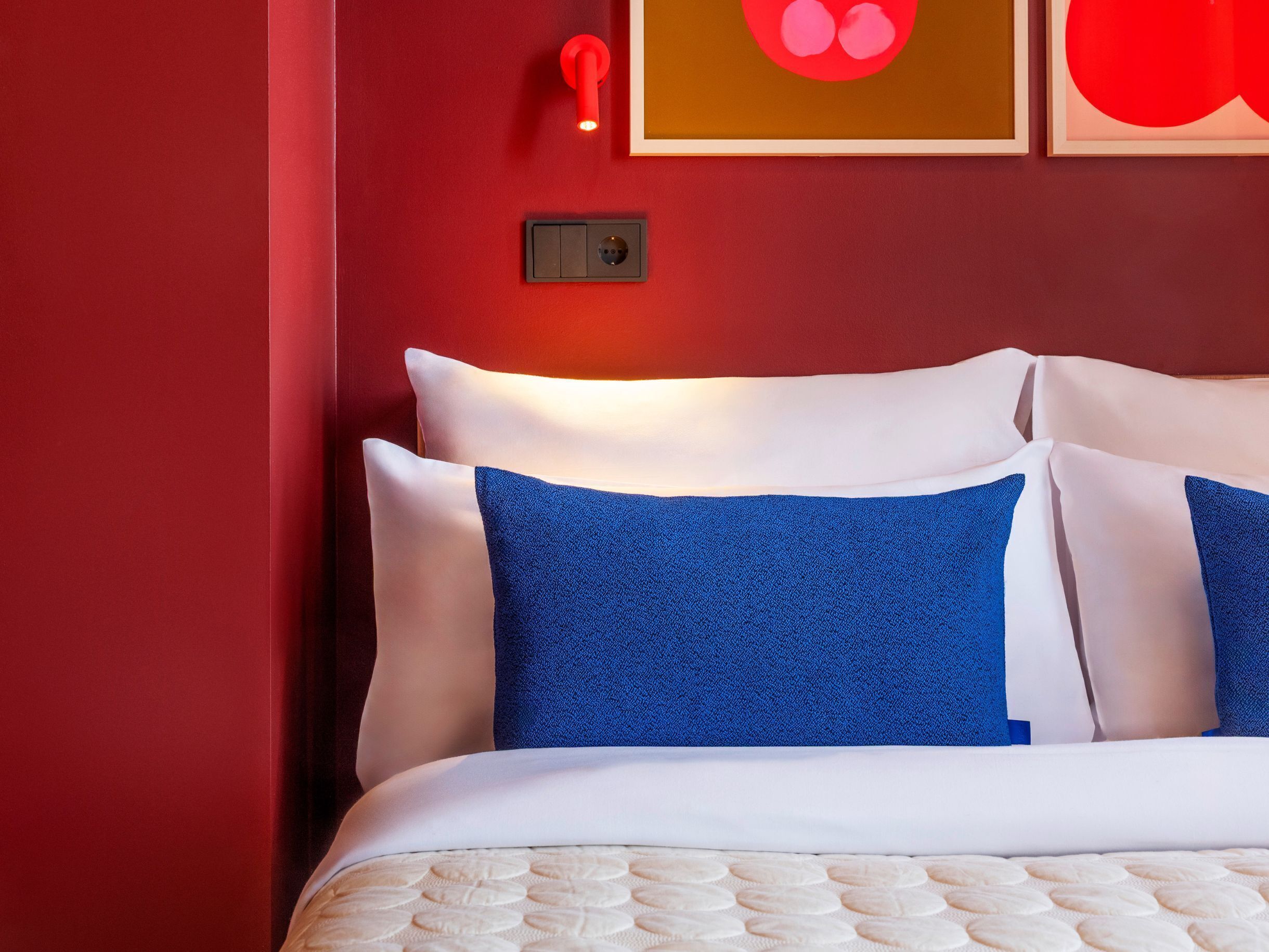 Un lit avec des oreillers blancs et bleus, un mur rouge et un interrupteur.