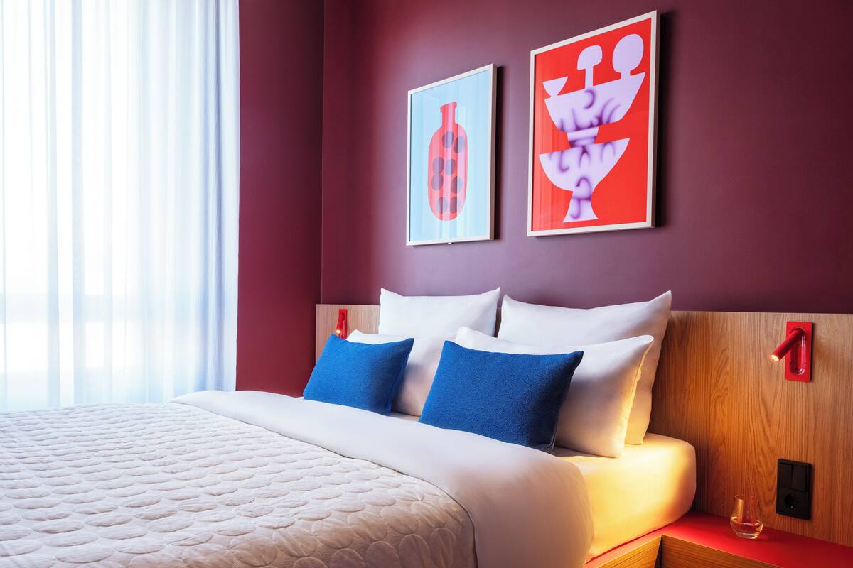 Una habitación de hotel con una cama grande, dos almohadas azules, un cabecero de madera y dos cuadros abstractos en la pared.
