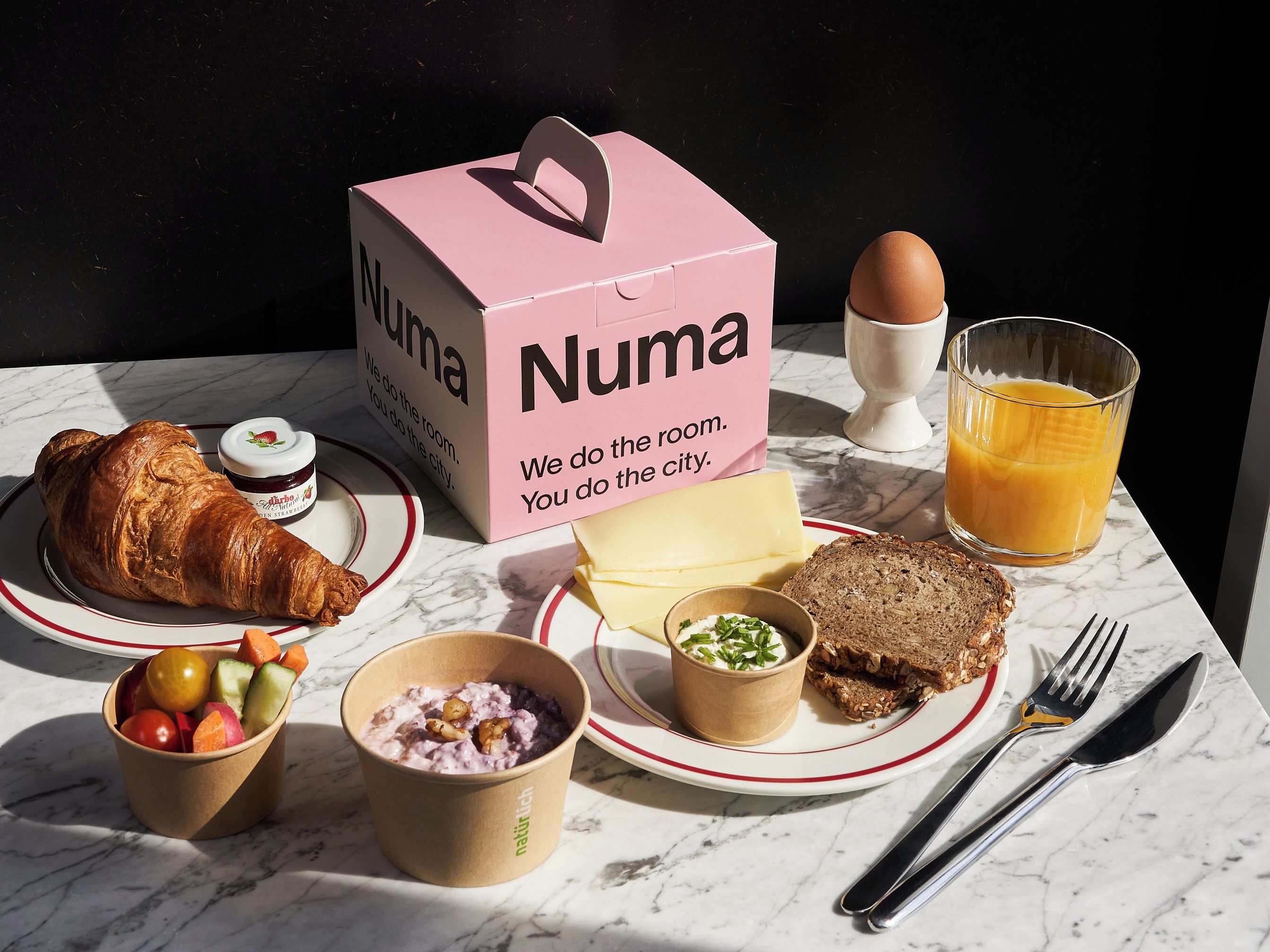 Un desayuno con un croissant, un vaso de jugo de naranja, un sándwich, un tazón de fruta, una taza de yogur y una caja con el nombre 'Numa'.