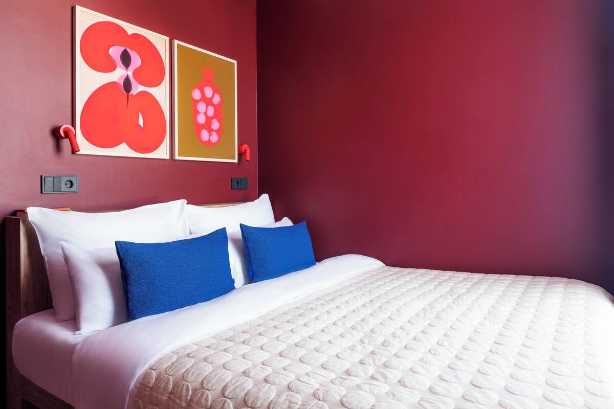 Una cama bien hecha con almohadas blancas y azules, un colcha color beige y dos cuadros abstractos en la pared.