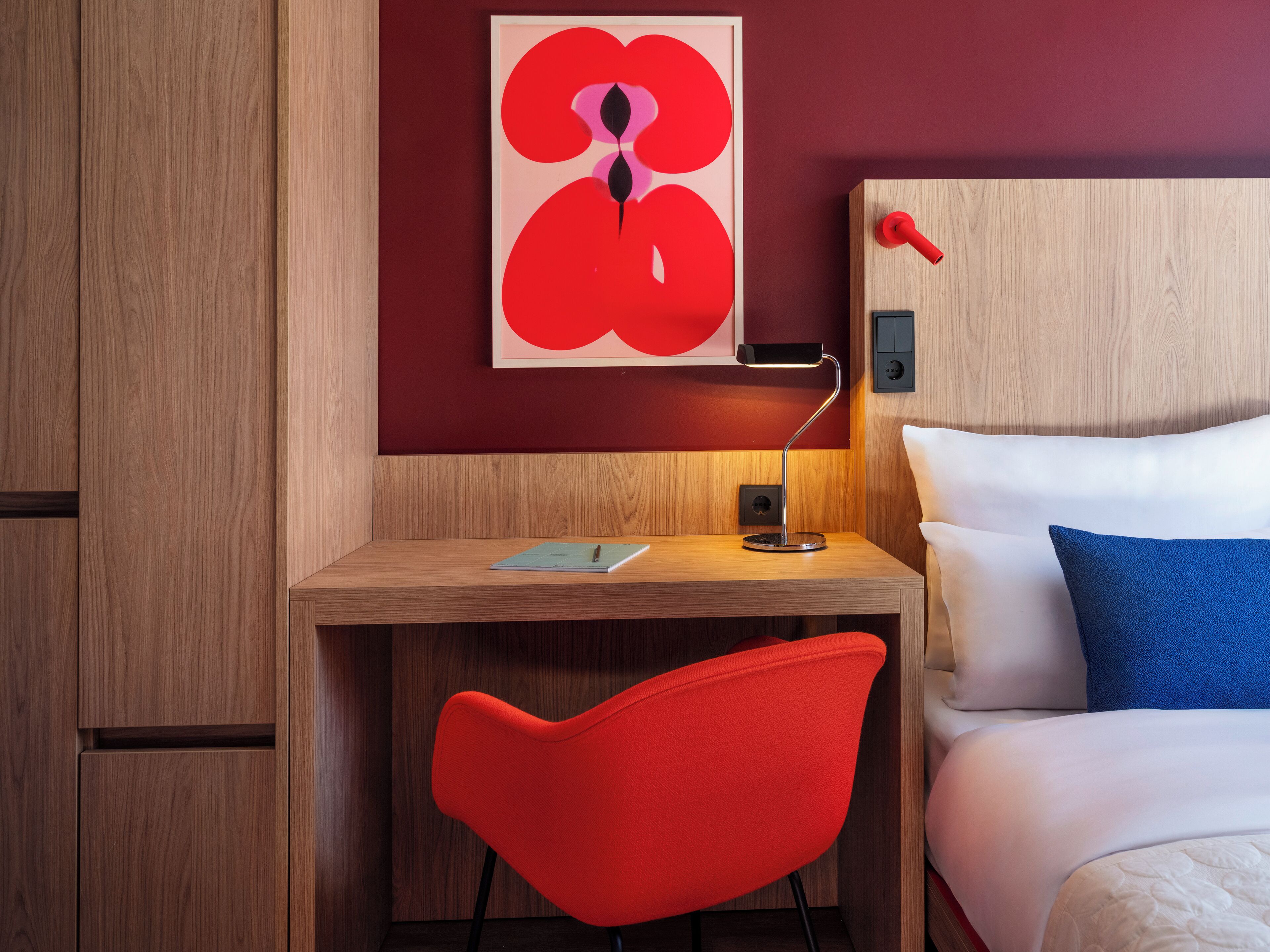 A hotel room with a wooden desk, a red chair, a bed with white and blue pillows, and a red abstract painting on the wall.