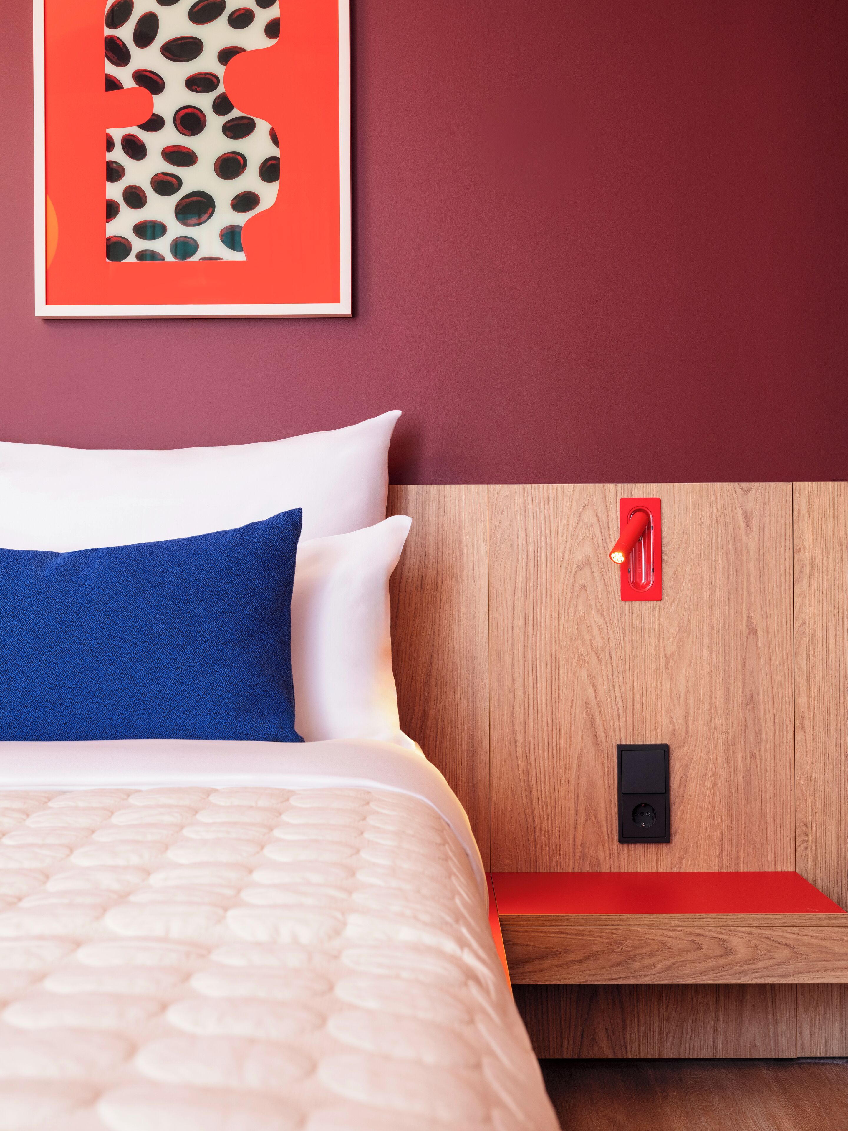 Un lit bien fait, avec un oreiller bleu, une tête de lit en bois, un objet rouge accroché au mur et un petit appareil noir posé sur la tête de lit.