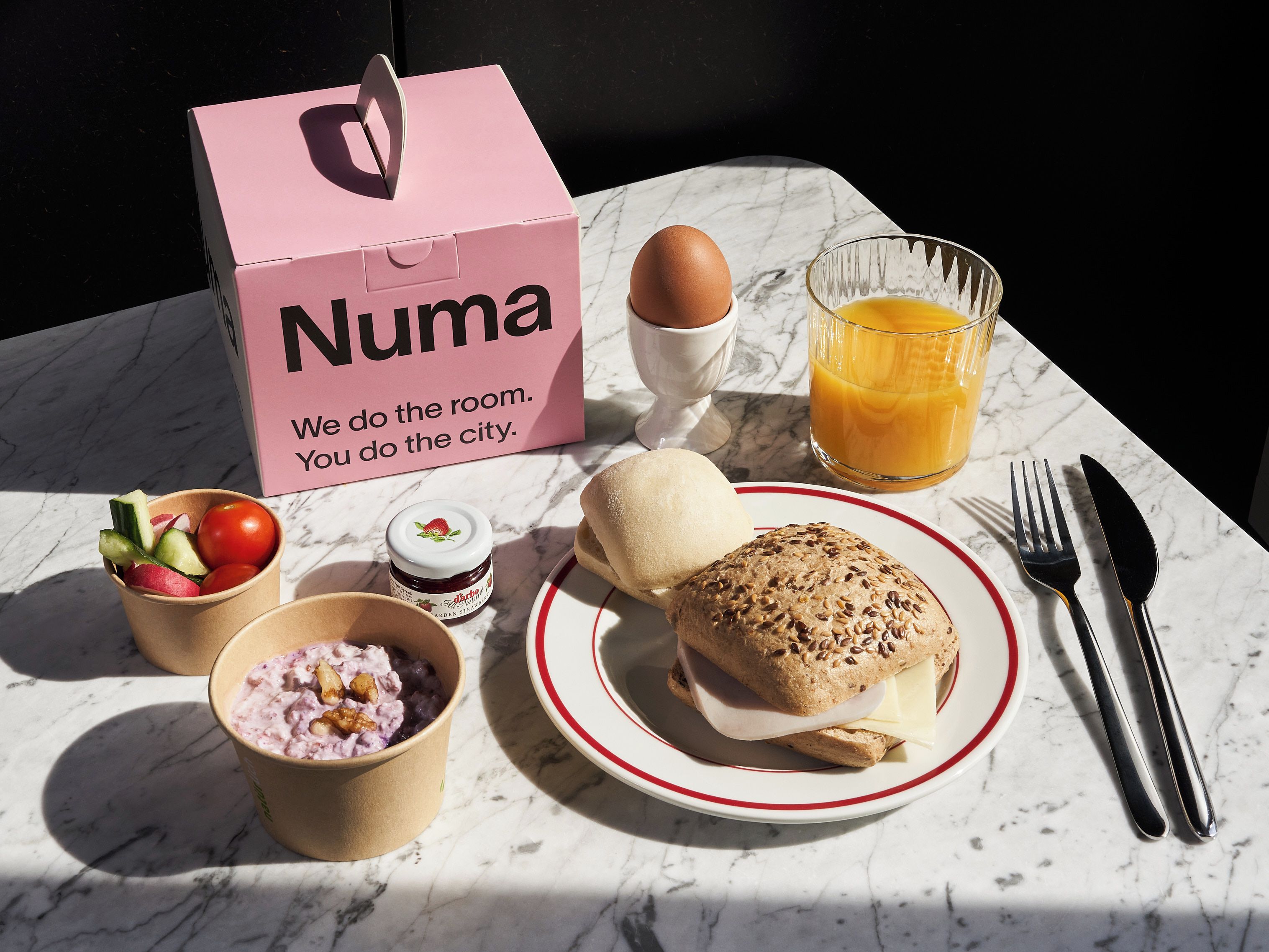 A meal setup with a sandwich, a glass of orange juice, a bowl of fruit, a cup of yogurt, and a box labeled 'Numa'.