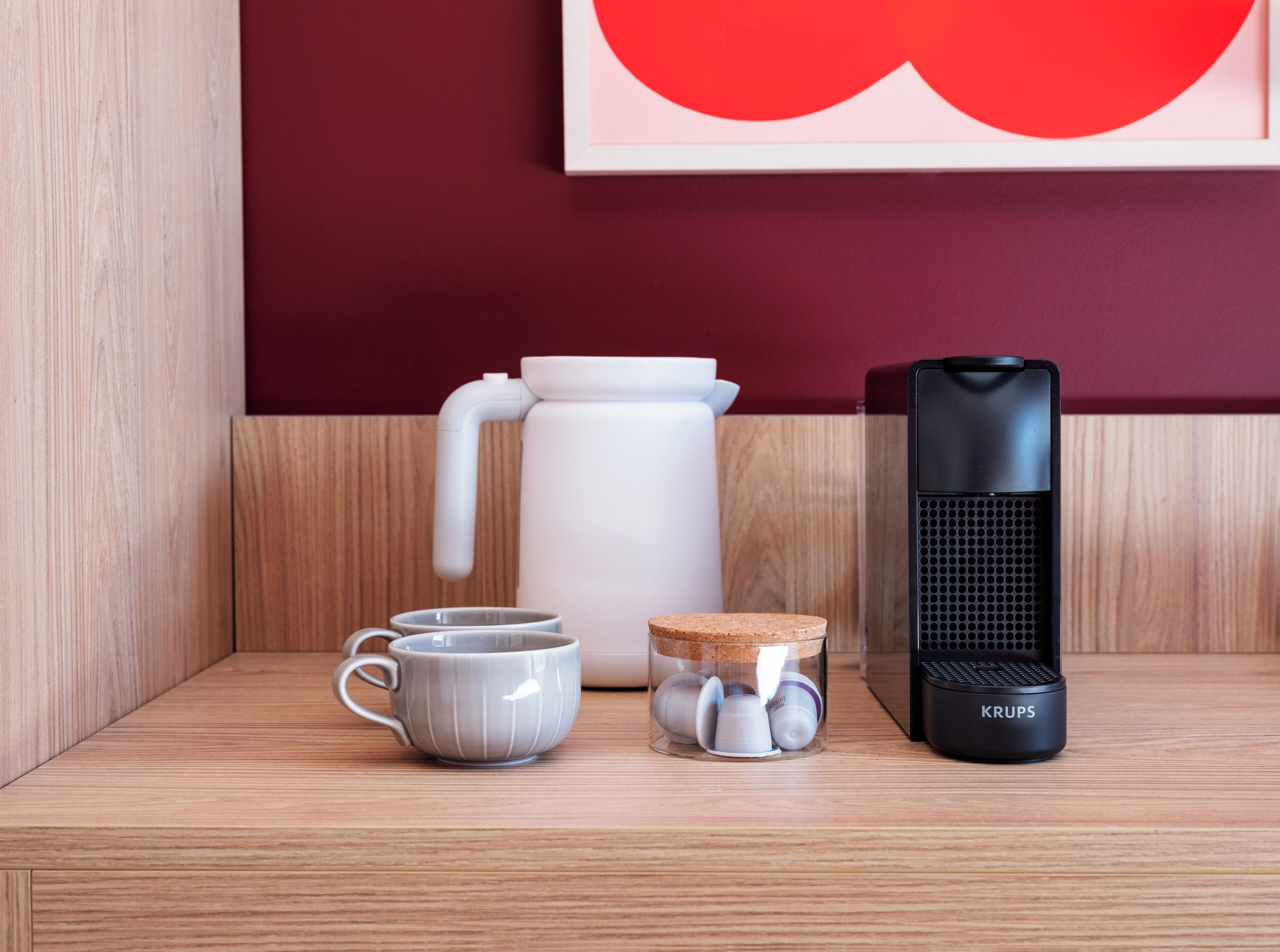 Une étagère en bois sur laquelle se trouvent une bouilloire blanche, deux tasses blanches, un bocal en verre muni de bouchons en liège et un petit radiateur électrique portable de la marque Krups, de couleur noire.