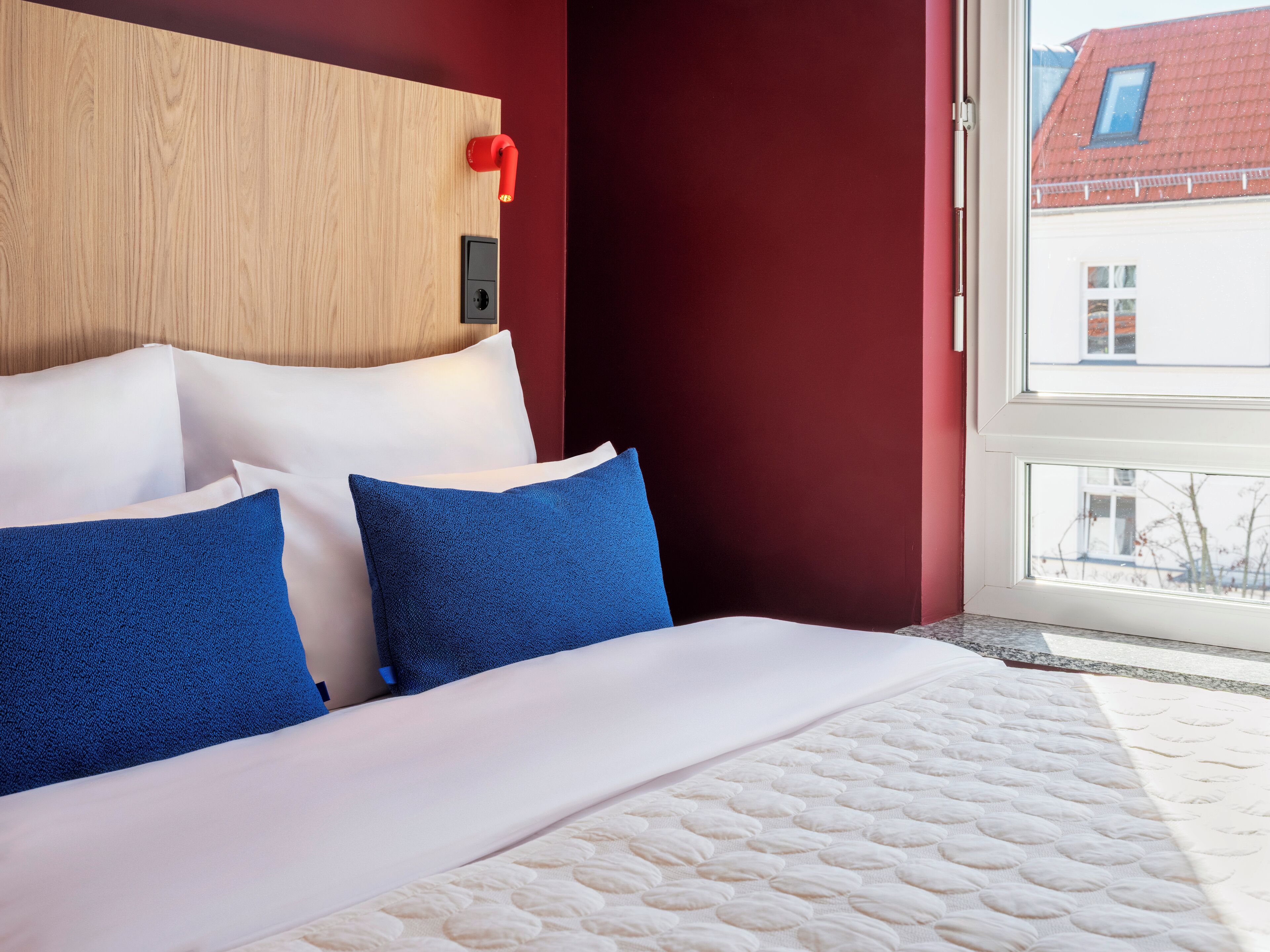 Ein Hotelzimmer mit einem hölzernen Kopfteil, weißer Bettwäsche und Blick auf Gebäude vor dem Fenster.