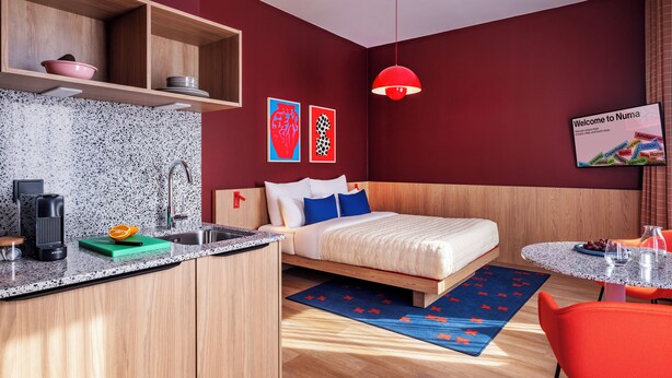Una habitación de hotel moderna con una cama de madera, una pequeña cocina, una lámpara colgante roja y un letrero de bienvenida.