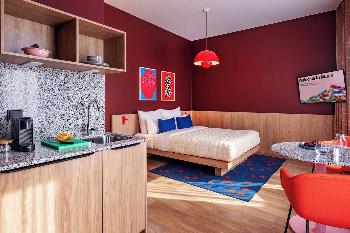 Una habitación de hotel moderna con una cama de madera, una pequeña cocina, una lámpara colgante roja y un letrero de bienvenida.
