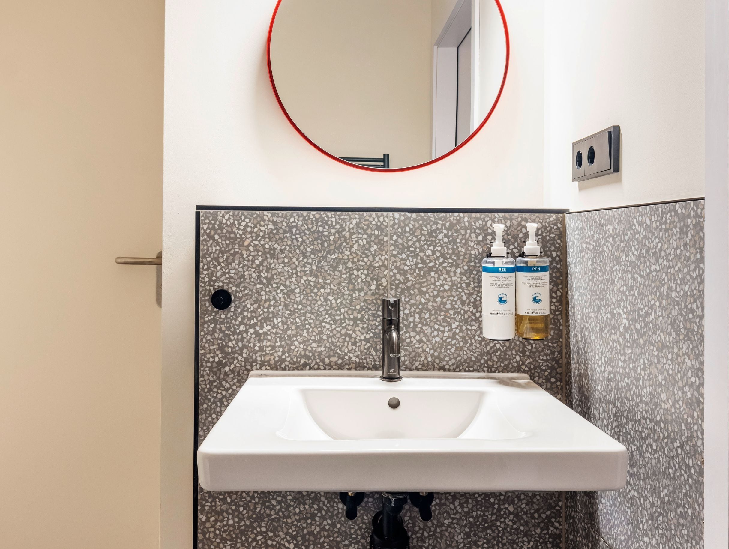 A bathroom with a white sink, a round mirror with a red frame, and two bottles of hand sanitizer on the wall.