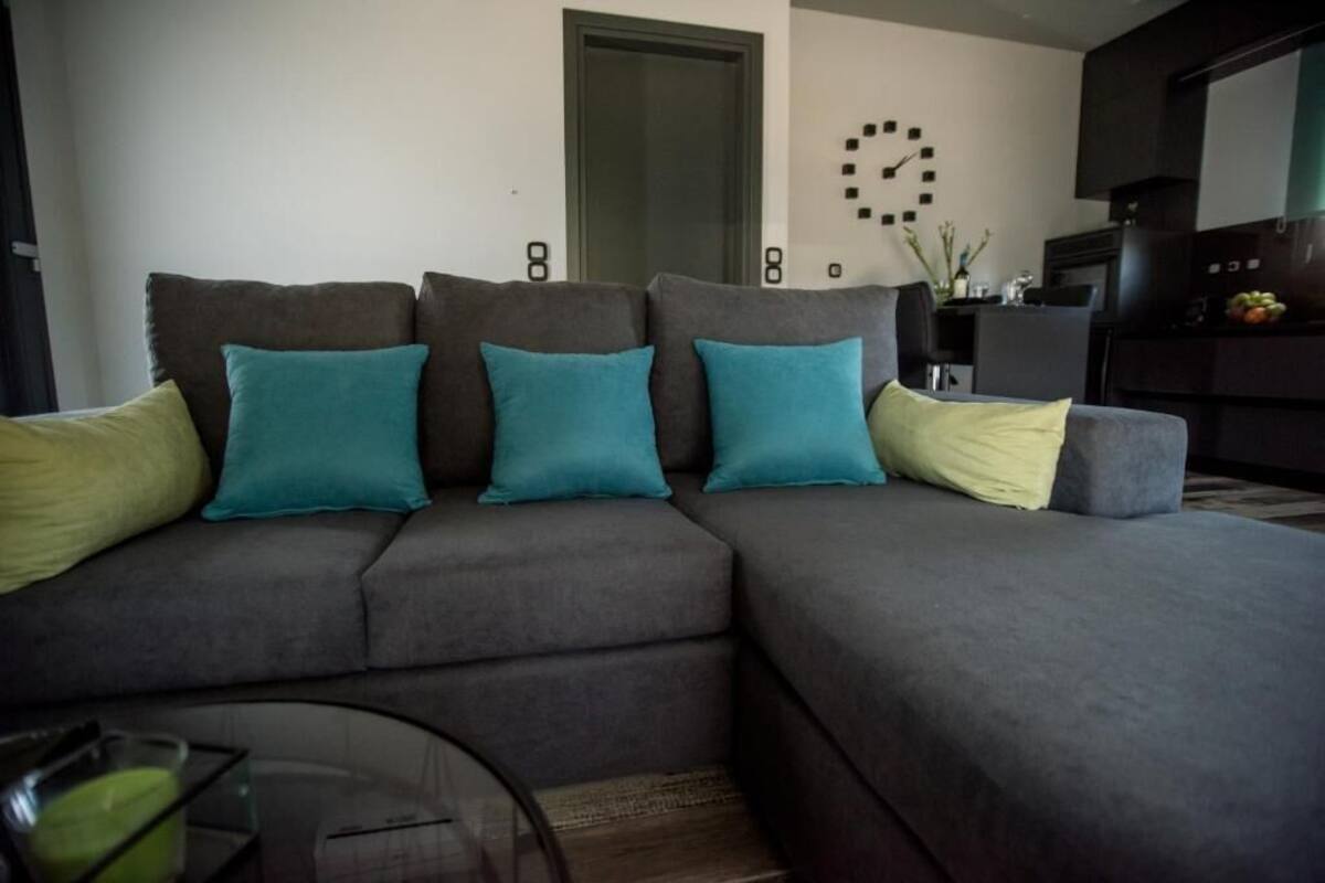 A modern living room with a grey sectional sofa, teal and yellow cushions, a glass coffee table, and a wall clock.