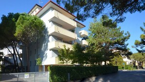 A multi-story residential building with balconies, surrounded by trees and a hedge. - Cozy apartment in a quiet area of Bibione (Bibione)