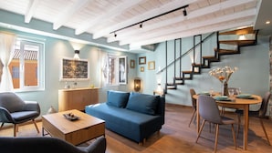 Un salon moderne avec un canapé bleu, une table basse en bois et un coin repas comprenant une table ronde et des chaises. Un escalier avec une rampe noire mène à l’étage supérieur.