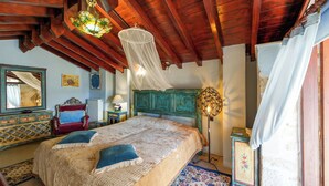 A bedroom with a wooden ceiling, a bed with a white and beige bedspread, a red chair, a blue sofa, a wooden cabinet, and a wall-mounted lamp.