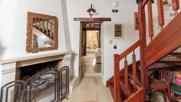A narrow hallway with a wooden staircase on the right, a fireplace on the left, and a door leading to a stone exterior.