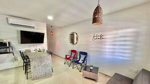 A modern living room with a grey sofa, a TV, and a kitchen area with a granite countertop and bar stools.