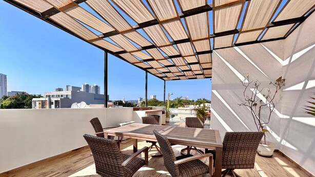 A rooftop terrace with a wooden table and wicker chairs, surrounded by a wooden floor and a metal frame structure.