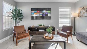 A modern living room with a flat-screen TV displaying streaming services, two leather armchairs, a coffee table with decorative items, and a grey sofa. - Rare 6BR Gem w/ Pickleball, Pool, & Arcade in NLV (North Las Vegas)