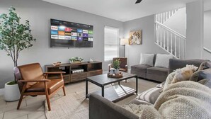 A modern living room with a flat-screen TV mounted on the wall, a ceiling fan, a sofa, a coffee table, and a staircase in the background. - Rare 6BR Gem w/ Pickleball, Pool, & Arcade in NLV (North Las Vegas)
