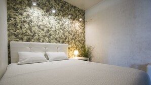 Un llit ben fet amb roba de llit i almoadines blanques, una paret amb disseny de marbre al fons i una petita taula de nit amb una làmpada i una planta.