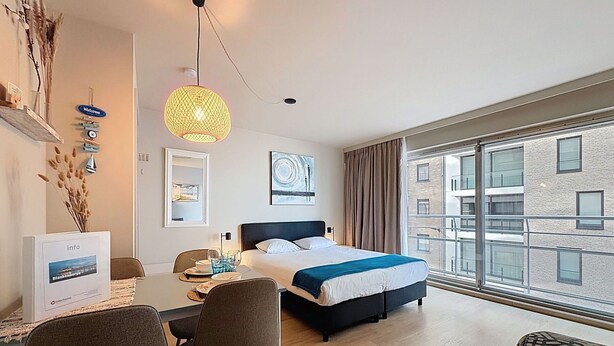 Una habitación de hotel moderna con cama, una mesa de comedor con sillas, un escritorio, una computadora y un amplio ventanal con vista a la ciudad.
