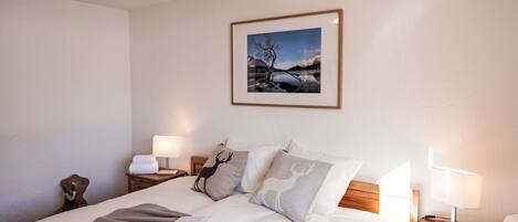 A neatly made bed with a striped blanket and a book, two bedside lamps, a framed picture of a landscape, and a nightstand with a lamp and a towel.