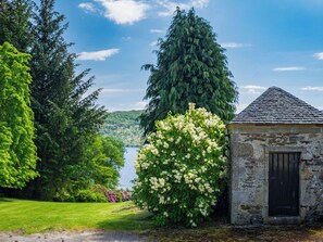 A stone building with a black door, surrounded by lush greenery and a view of a lake.
