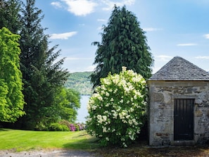 A stone building with a black door, surrounded by lush greenery and a view of a lake.
