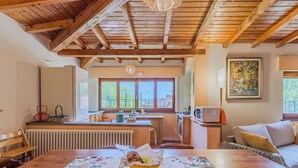 Une cuisine chaleureuse avec des poutres en bois, une table à manger agrémentée d’un chemin de table à motifs floraux et une vue sur l’extérieur.