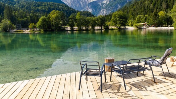 A wooden deck with metal chairs and a table overlooking a clear lake with mountains in the background.