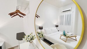 Une chambre d’hôtel moderne avec un grand miroir rond, un lit avec du linge de maison blanc, une table de chevet avec une lampe, une petite table de chevet avec un plateau, et un climatiseur fixé au mur.