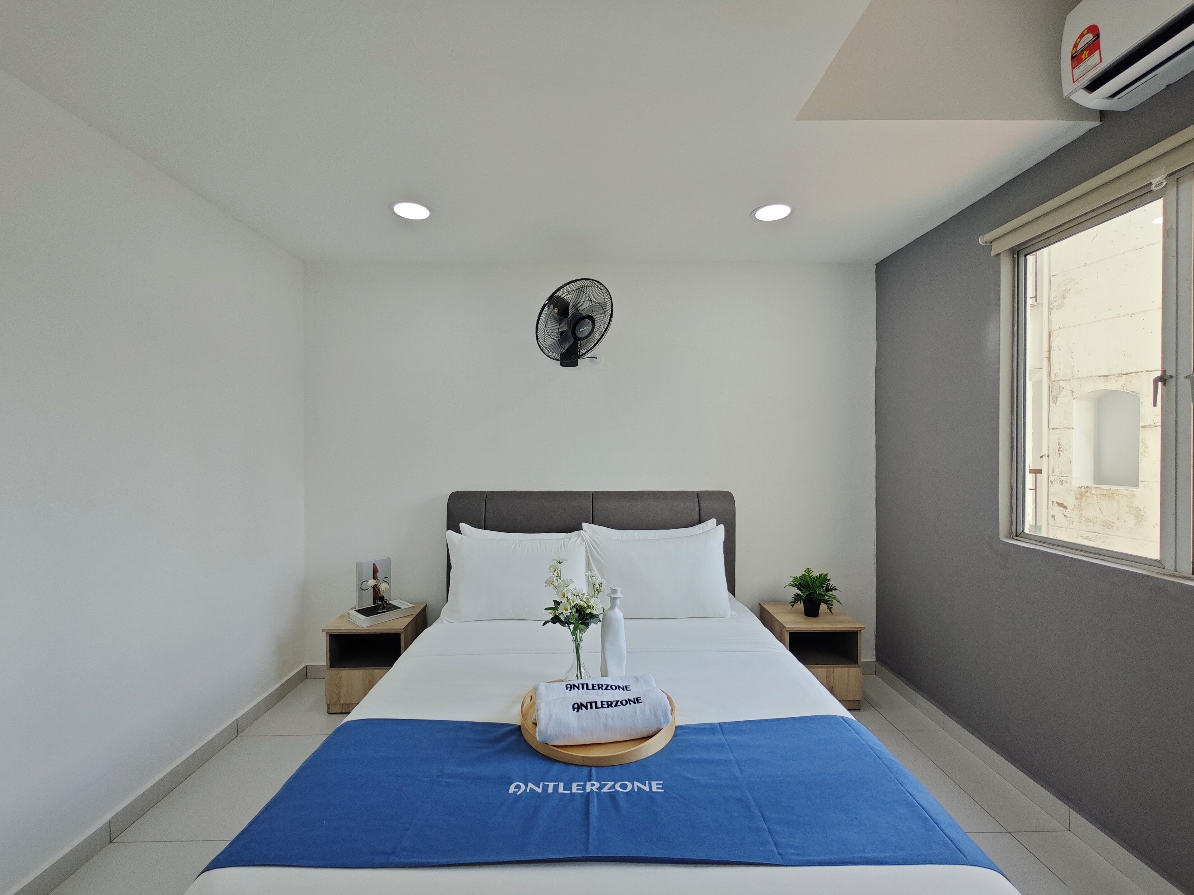A hotel room with a bed, a bedside table with a lamp, a wall-mounted fan, and a window with a view.