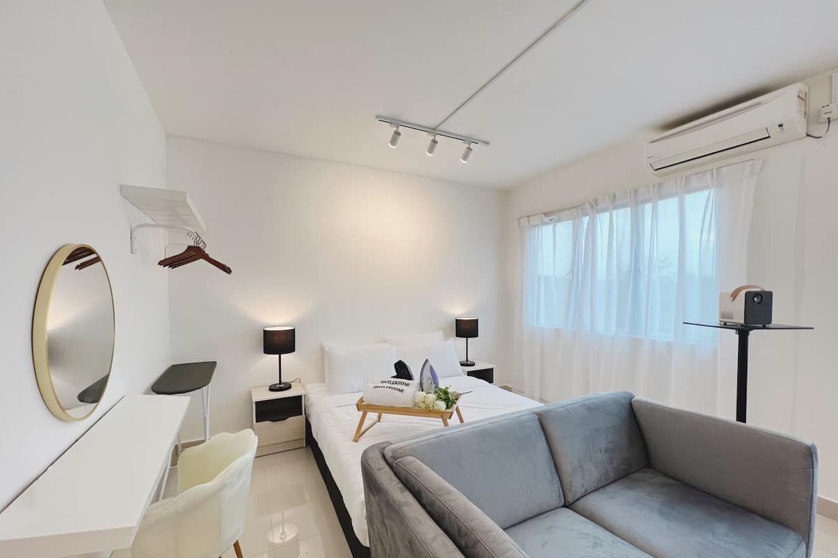 Une chambre d’hôtel moderne comprenant un lit, un canapé gris, un bureau, une chaise, une table de chevet, une lampe, un miroir et une fenêtre avec des rideaux.