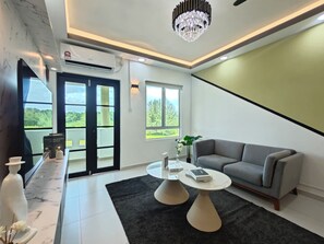 Un salon moderne avec un canapé gris, une table basse blanche et un tapis noir. Une grande fenêtre offre une vue sur la verdure à l’extérieur.