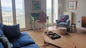 Un salon spacieux avec une grande fenêtre, un bureau vert, un fauteuil bleu à motifs et une table basse en bois.