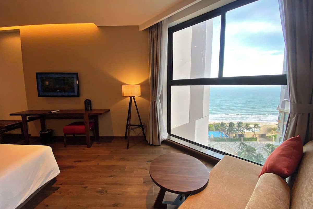 A hotel room with a bed, a desk, a round table, a sofa, and a large window with a view of the ocean.