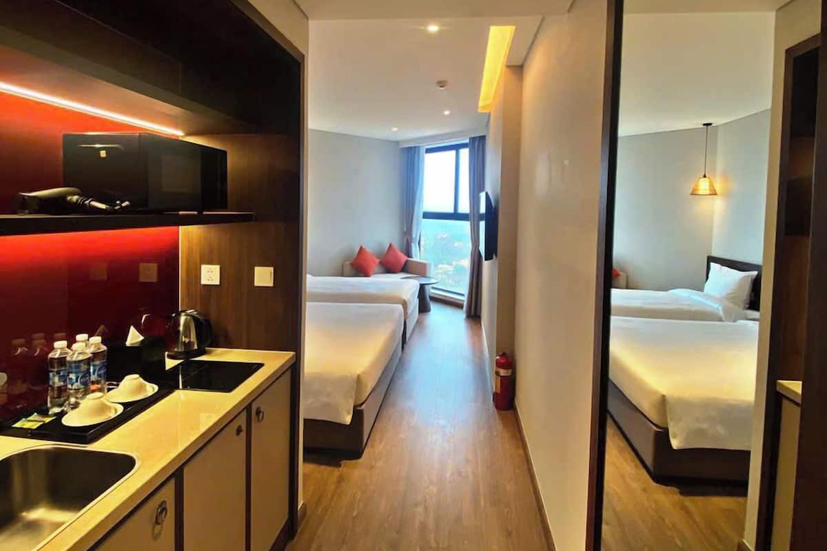 A hotel room with a kitchenette, two beds, a window with a view, and a mirror reflecting the room.