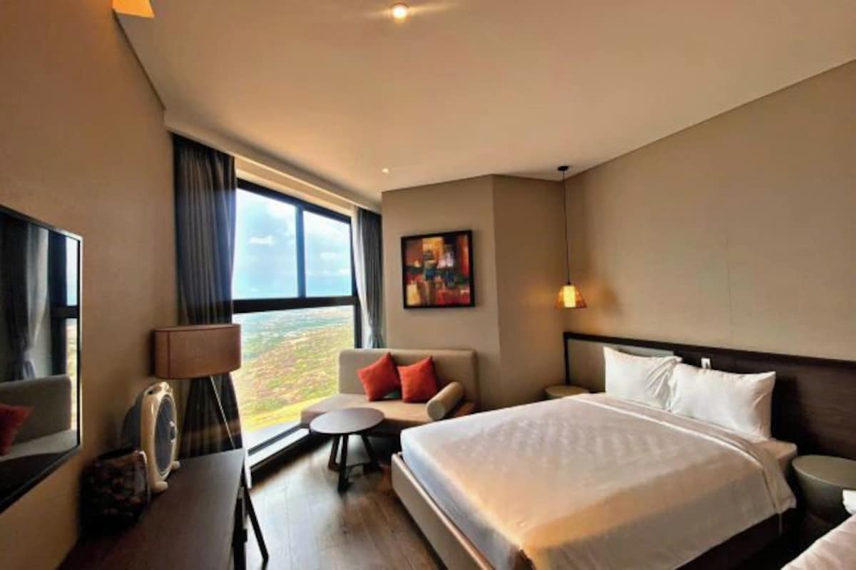 Habitación de hotel con una cama grande, un sofá, una mesita, una lámpara y un amplio ventanal con vistas.