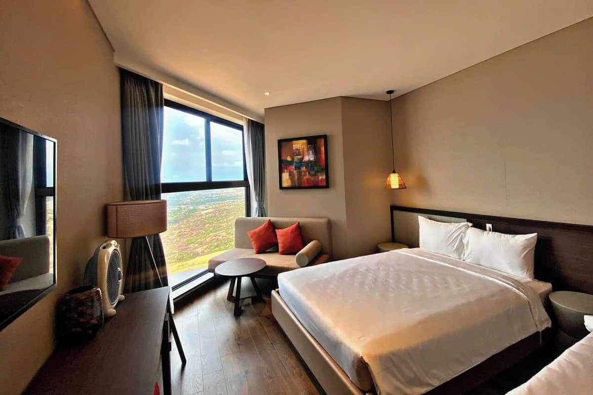 A hotel room with a large bed, a sofa, a small table, a lamp, a TV, and a window with a view.