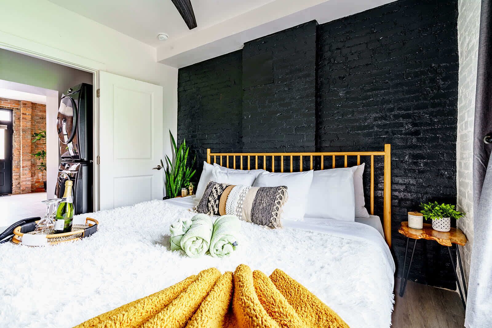 Un dormitorio moderno con pared de ladrillo negro, estructura de cama de madera, ropa de cama blanca y una mesa para el desayuno con pan y una botella de champán.