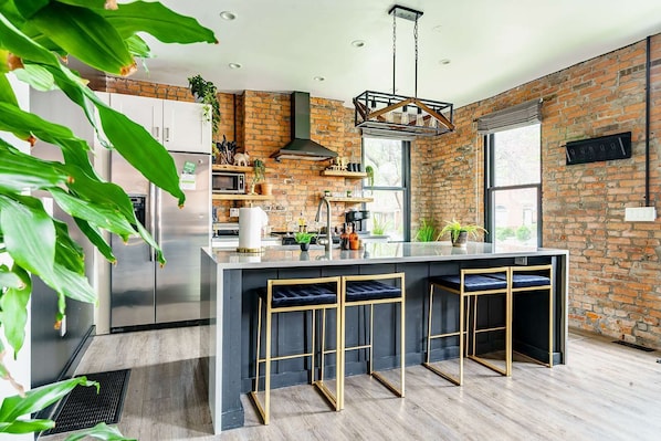 A modern kitchen with a central island, bar stools, a hanging light fixture, and a brick wall.