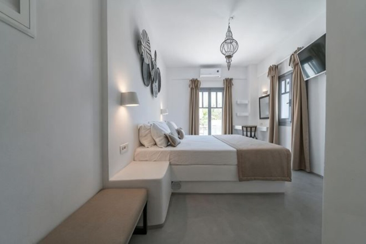 Un dormitorio con cama, mesitas de noche, un banco, una lámpara colgante y una ventana con cortinas.