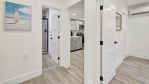 A modern kitchen with white cabinets and a central island, a hallway with a glass door leading to a bathroom, and a dining area with a table and chairs. - Banyan St 2BR/1BA off Las Olas- Steps to Beach #1 (Fort Lauderdale)