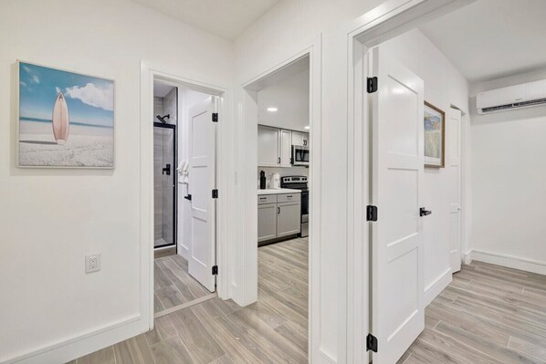 Una cocina moderna con armarios blancos y una isla central, un pasillo con una puerta de vidrio que da a un baño, y un comedor con una mesa y sillas.