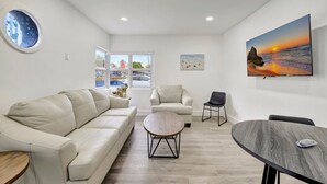A modern living room with a beige sofa, a round coffee table, and a dining table with chairs. There is a painting of a sunset on the wall and a window with a view of the outside. - Banyan St 2BR/1BA off Las Olas- Steps to Beach #1 (Fort Lauderdale)