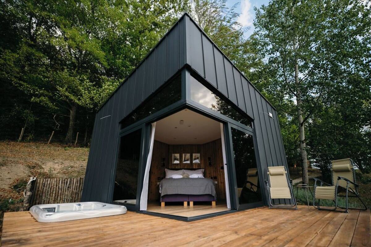 Una cabaña moderna, de color oscuro, con un amplio ventanal que da a un dormitorio con una cama y dos sillas.