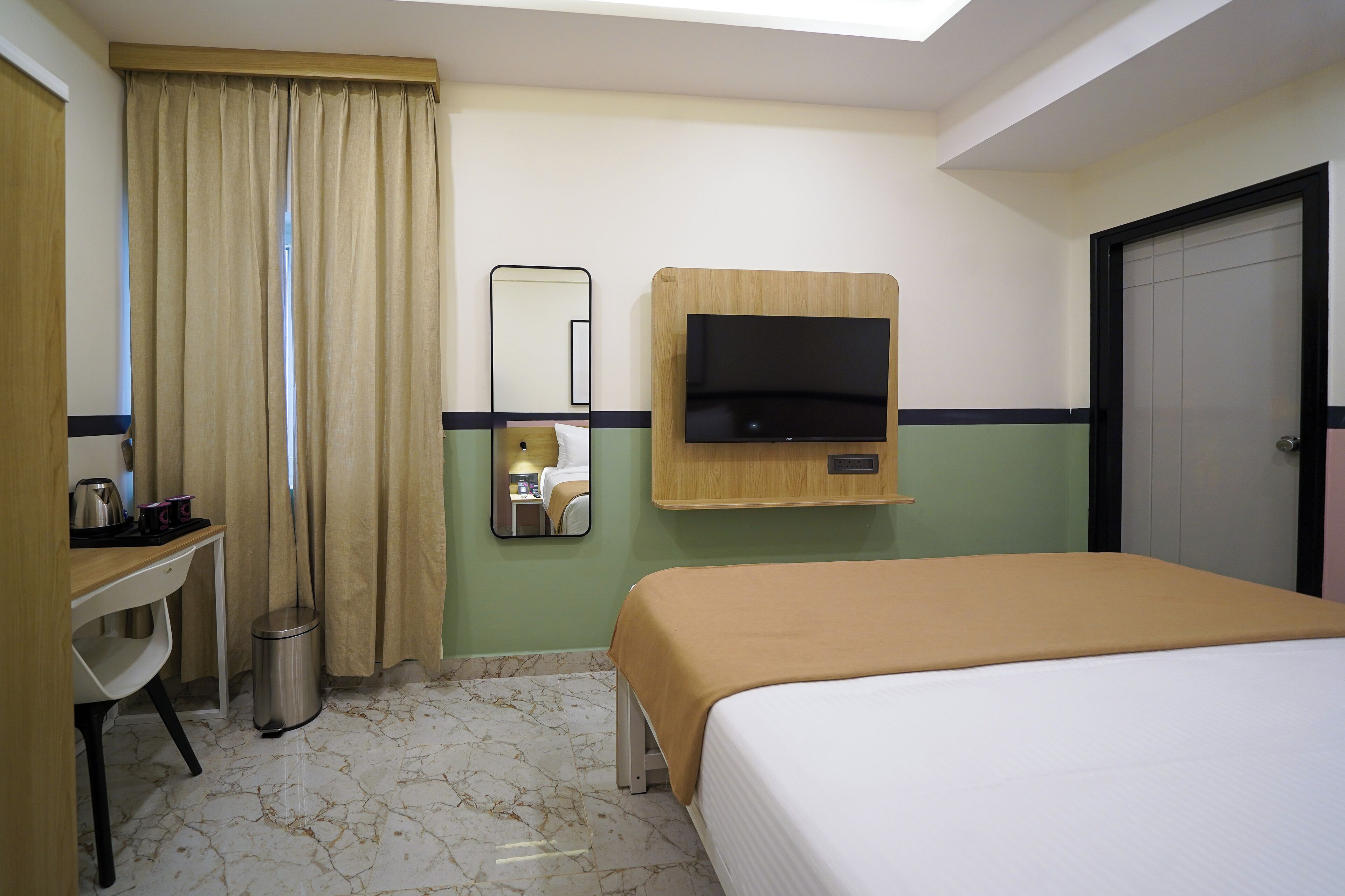 A hotel room with a bed, a TV mounted on the wall, a desk with a chair, a trash bin, and a door.