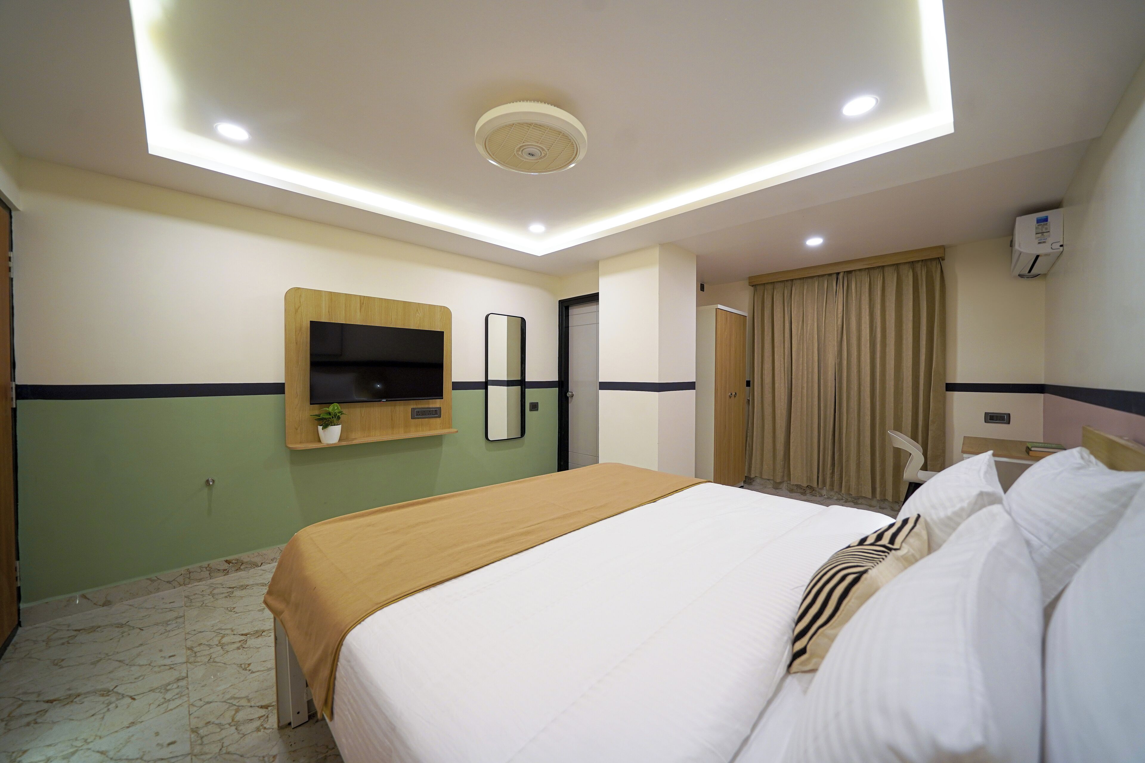A hotel room with a large bed, a flat-screen TV mounted on the wall, a bedside table with a lamp, and a window with curtains.