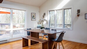Un comedor con mesa de madera, sillas y un banco. Amplias ventanas con vista a árboles y una terraza exterior.