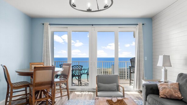 A room with a dining table, chairs, a sofa, a lamp, and a view of the ocean through large sliding doors.
