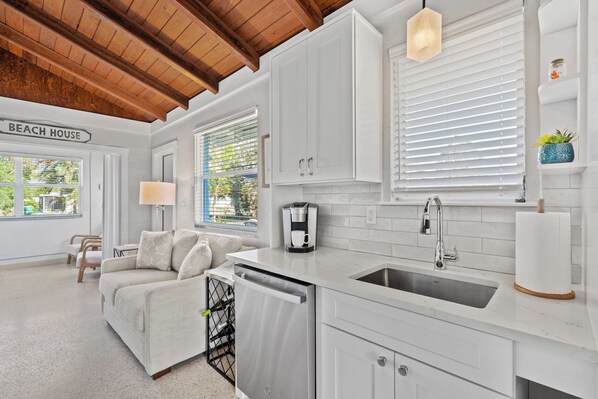 A modern kitchen with white cabinetry, a dishwasher, and a sink. There is a sign that reads "BEACH HOUSE" on the wall.