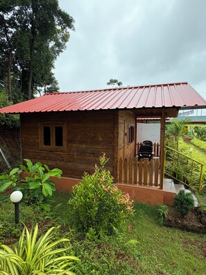 A wooden cabin with a red roof, a porch with a wooden fence, and a garden with various plants. - Hill View Paradise (Gokarna)